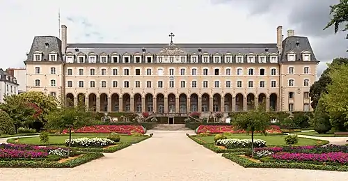 A large four-storey sand-coloured building with 14 arches at the ground-floor entrance and a large landscaped formal garden in front.  The photograph was taken during the summer, so many flowers are in bloom and there are leaves on the bushes and trees.