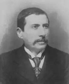 Black-and-white photograph of a white man, in middle age, with short black hair and a moustache. He wears a jacket, collared shirt and tie, and faces slightly to the camera's right.