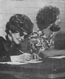 Photo of Woods sitting next to a woman who is writing on a piece of paper. Woods is facing away from the camera. He has curly hair and a beard and is wearing a short-sleeved floral shirt.