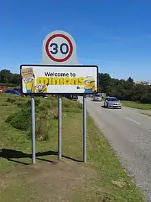 road sign (tempo limit 30): Welcome to minions; the word "minions" is written in the font from the film, and surrounded by three "minions", one of whom is shown holding a cardboard sign that reads "Please drive carefully"