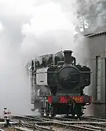 A green pannier tank locomotive appears from a cloud of its own steam and smoke.