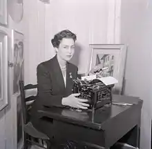 A fair-skinned woman with dark coiffed hair, wearing a dark suit and seated at a small desk with a manual typewriter