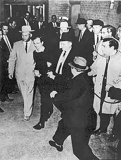 A black-and-white photograph showing a man with men on either side of him walking past people on either side, from above. At the bottom right a man with a hat has a gun pointed at the man in the center.