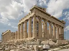 Ruins of a white temple with columns on a rock above a city.