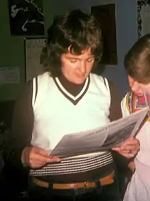 Color photograph of a short-haired woman wearing a white sweater vest over a brown top and reading a newspaper. To her left is another woman reading the same paper.