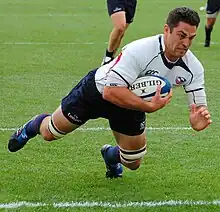 A man wearing a rugby uniform consisting of a white shirt, dark blue shorts, and dark blue socks dives in an attempt to score a try.