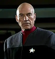 A film-screenshot of a balding Caucasoid man in a science-fiction military uniform; he is facing the camera, and looking slightly to its right