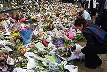 A woman adds a flower arrangement to a large memorial display set against a fence.