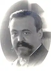 close-up photograph of a youngish white man with full head of neat dark hair and a large moustache and neat short beard