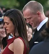 A brown haired woman signs autographs for fans. She wears a red dress. Behind her there is a blond man in a suit. The woman and the man are facing a crowd of fans.