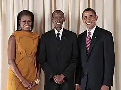 Michelle Obama, Paul Kagame and Barack Obama, standing and smiling in front of a curtain
