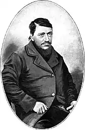 A confident-looking man of about 40 with a large dark beard. The thumb on his left hand is absent.