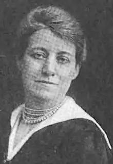 A middle-aged white woman, wearing several strands of beads and a white collar over a black dress.