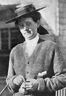 White woman standing outside, wearing a small straw hat and a cardigan.