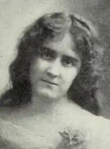 A young white woman with long loose dark hair, wearing a white lacy dress with an open neckline