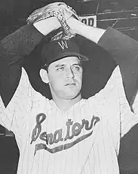 A man in a white baseball uniform with dark pinstripes with "Senators" on the chest wearing a dark cap holds his hands above his head in a baseball glove