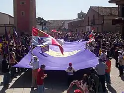 People at a celebration holding a huge purple flag.  Others wave different flags, such as the Cross of Burgundy or the modern flag of Castile and León.
