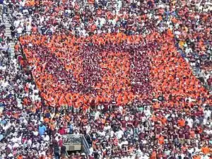 People in the stadium wearing orange and maroon T-shirts to form an image of the letters "VT" in maroon on a square background of orange.