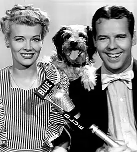 Penny Singleton as Blondie and Arthur Lake as Dagwood Bumstead, from a 1944 publicity photograph: A smiling white woman with blonde curls and a striped pinafore apron; a smiling white man wearing a bow tie; a small dog posed between their shoulders; and a microphone in front of them, labeled "Blue"
