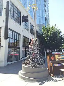 Photograph of a curved pole topped with a gold-colored small bicycle; at the base is a collection of small bicycles