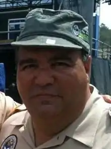 A Hispanic male wearing a beige colored button-up shirt and green cap posing for a picture outdoors.