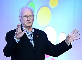 A photograph of Pete Waterman, an elderly Caucasian man with white hair, wearing a suit, sitting with his arms folded