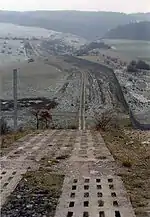 A patrol road, made of two parallel rows of perforated concrete blocks, performs a steep descent into a valley. To the right, running parallel to the road, is a continuous fence. The road and fence continue into the distance, crossing fields that are dusted with a light sprinkling of snow, and ascending another hillside on the far side of valley. Dark forests loom in the distance.