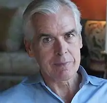 An older white man with a blue collared shirt is looking directly into the camera with a small smile on his face.