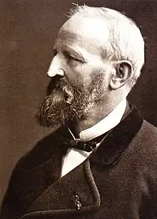 A Caucasian man in his 60s with thinning white hair and a greying moustache and beard, wearing a white shirt with an upturned collar, a thin dark bow tie, and a jacket of heavy material. He is facing left.