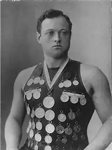 A man aged 19 with short hair looking off to one side. He wears a vest with medals pinned to it, along with one medal around his neck.