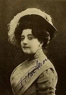 A middle-aged white woman wearing a large plumed hat and a corseted dress with a high collar. Her dark hair is in an updo. She is not smiling, but looking at the camera sidelong.