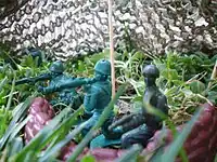 Three green plastic bazooka men on dirt in between several strands of grass