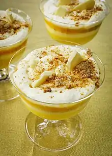 A close up image of a Platinum Jubilee pudding, taken from an event at the British Library in May 2022. It focusses on a white cream top with Amaretti crumb, over a yellow toned layered trifle. Shards of white chocolate are also on top of the cream.