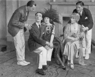 Two young white men stand crouched next to three white seated people; all are in early 1920s costumes