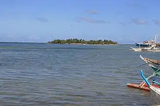 Small low green island in blue sea, at about 600 m, with several palm trees on top.  To right prows of four bangkas – local boats for fishing or diving.