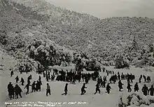 Several dozen students move around a clearing on a forested mountainside with approximately a foot of recent snowfall