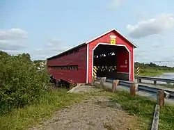 Single-track bridge over a river in a marshy area with a wooden enclosure with flaking red pain; there are signs which say "1930 Molesworth", "3,0m", and "baignade á vos risques" as well as yellow and black striped graphics at the entrance.