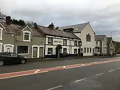 Main A470 road in foreground of black tarmac with red central stripe having residential buildings, pub and Memorial Hall stretched behind into distance away from camera position
