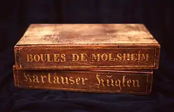 Two wooden boxes on top of each other, on which are written "Boules de Molsheim" and "Kartäuser Küglen" respectively.