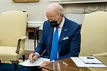 President Joe Biden is sitting on the yellow chair wearing a mask and blue suit, signing the Defense Production Act and launching Operation Fly Formula.