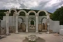 Five marble columns near the walls of a partially preserved building. A gate and a small monument are in the background.