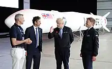 four men stand in front of a small space rocket, positioned horizontally, inside a hangar