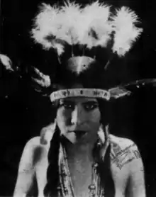 A young woman with fair skin and dark hair dressed in two braids, wearing a feathered headdress with a band across her brow