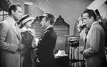 Black-and-white film screenshot of several people in a nightclub. A man on the far left is wearing a suit and has a woman standing next to him wearing a hat and dress. A man at the center is looking at the man on the left. A man on the far right is wearing a suit and looking at the other people.