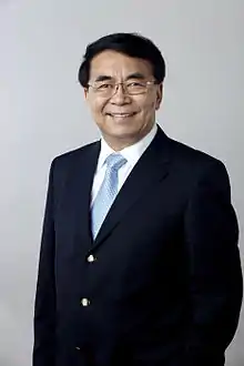 a smiling, aged man, wearing glasses, a suit and a blue tie while looking straight into the camera