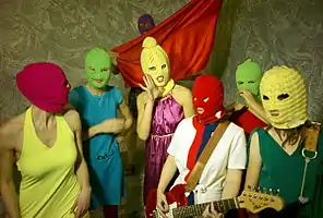 7 women with bright colored clothes and multicolored knit ski masks over their faces. A woman at the center holds a guitar and one at the back holds a piece of red fabric.