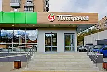 The front of a store, with glass windows and glass doors, and the name of the store in Russian on top, including a logo that appears to be the number five with a leave instead of the upper part of the number.