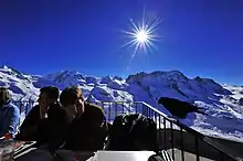  Visitors sitting at tables on a large balcony high in the Swiss Alps, and a chough is perching on a railing beside them.