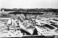 A large ship inside a dry dock. The dry dock is surrounded by industrial buildings and hills are visible in the background
