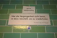 A green brick wall with a white sign reading "Wer die Vergangenheit nicht kennt, / ist dazu verurteilt, sie zu wiederholden. / (G. Santayana 1863–1953, Philosoph)